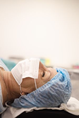 Woman in hair net and facemask lying down pre-surgery, vertical