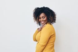 Studio shot of a smiling Black woman in yellow shirt with her hand on her shoulder and eyes closed