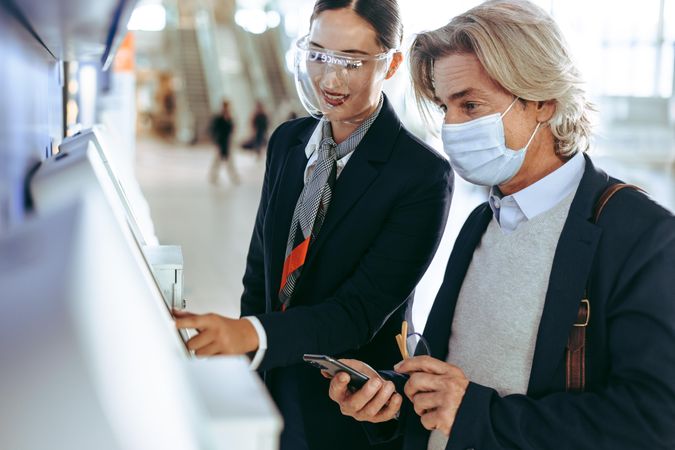 Masked businessman at self service check in machine with flight attendant