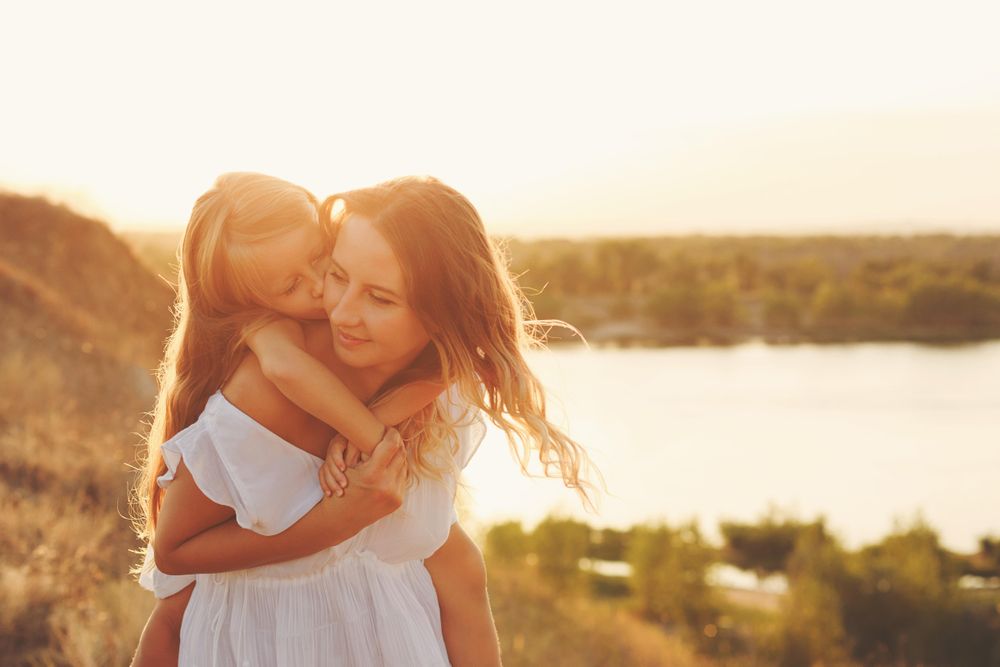 Young girl on woman’s back in front of river at dusk