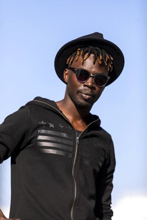 Black man wearing hat & sunglasses standing against blue sky