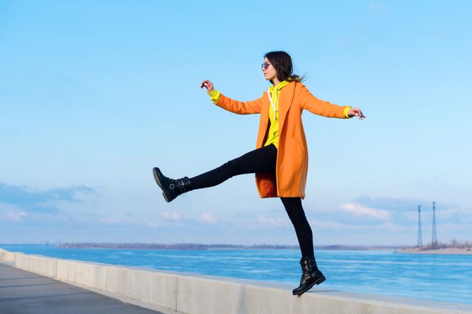 Woman in sunglasses, orange coat, yellow hoodie and jeans against the sea