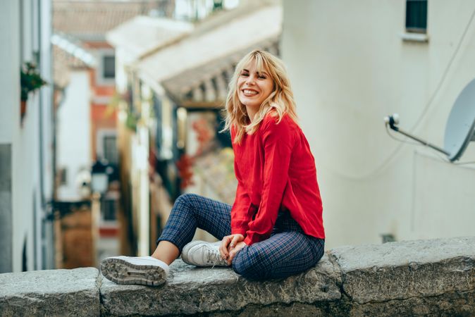 Smiling blond woman sitting on stone wall with legs crossed above Spanish street