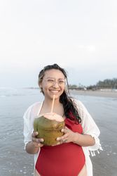 A young Hispanic woman is posing with a coconut on the sea edge at the beach