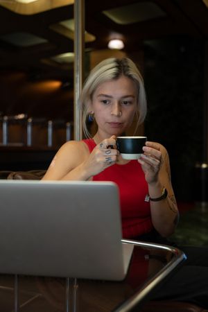Spanish student with a tattoo works on a laptop and drinks coffee to cheer up, sometimes looks at the clock that has time to finish work