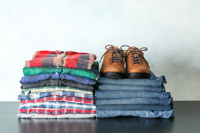 Stack of colorful shirts, jeans and shoes on table against light background, space for text