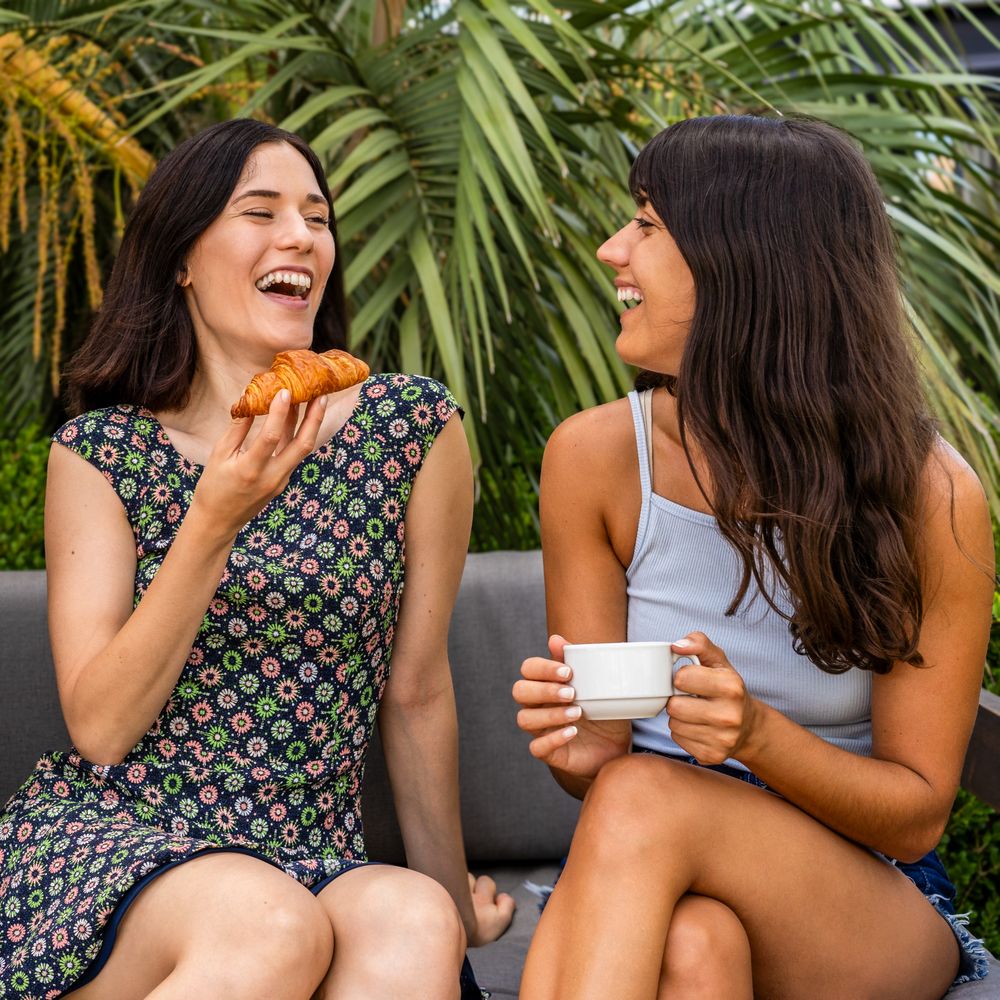 Two women are sitting on a couch talking and laughing, one of them is holding a croissant