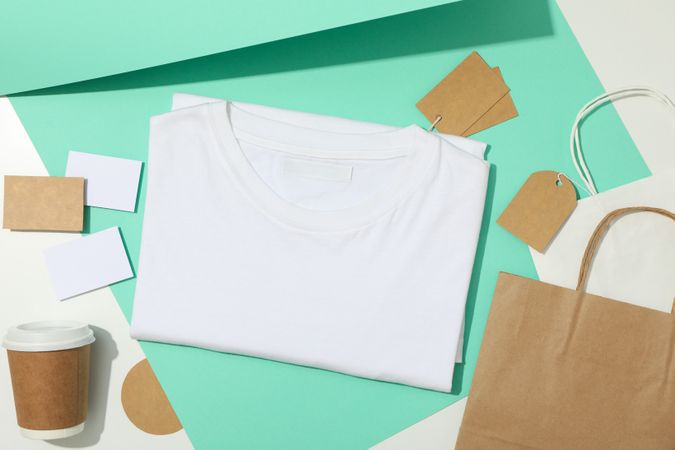Blank t-shirt, tags, paper cup and paper bags on mint and light background, space for text
