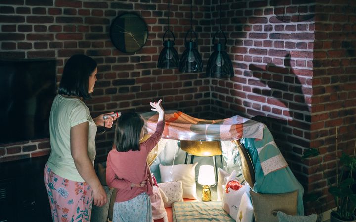 Back view of mother holding flashlight while daughter making shadow puppets with hands over wall.