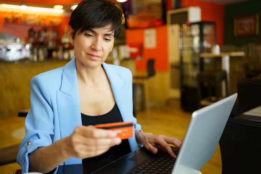 Female in trendy blue jacket sitting in cafe with laptop and looking at her credit card