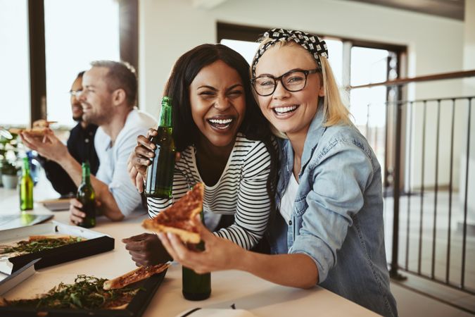 Two female colleagues smiling for the camera during a work lunch with pizza and beer