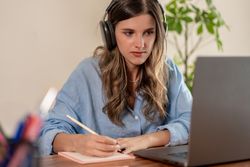 Focused young woman wearing headphones writing notes while working remotely at home