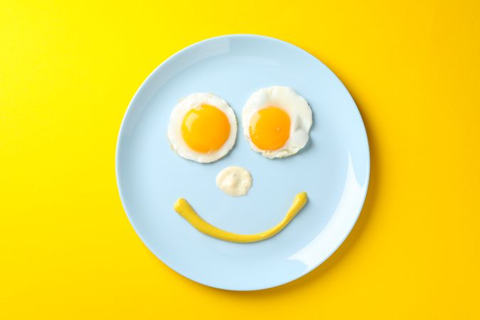 Looking down at blue plate on yellow table with smiley face on it made of eggs and condiments