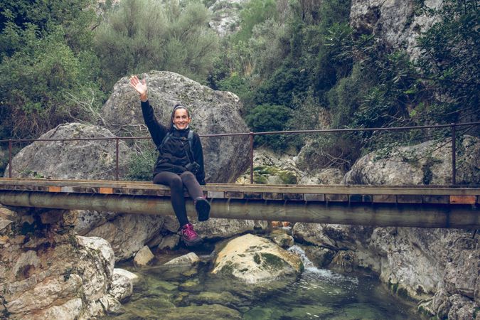 Delighted woman showing greeting gesture sitting on bridge
