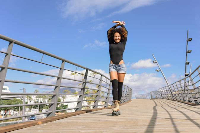 Woman with afro hairstyle in roller skates on wooden bridge with arms above head