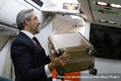 Male in business attire putting small suitcase above seats in airplane cabin bEqgob