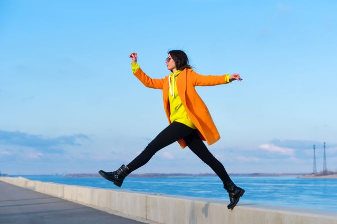 Woman in sunglasses, orange coat, yellow hoodie and jeans against the coastal background