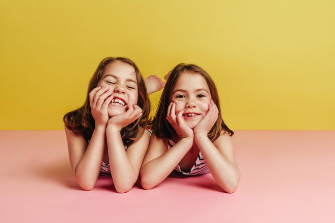 Twin girls lying on pink floor with hands on chin and looking at camera over yellow background