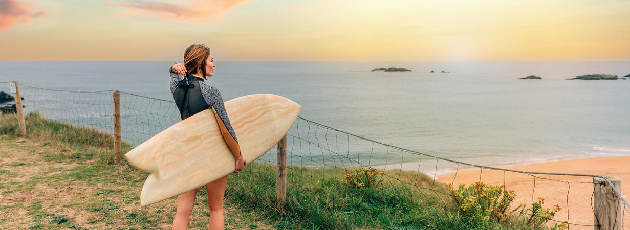 Female walking with surfboard atop of cliff with beautiful coastal view, banner