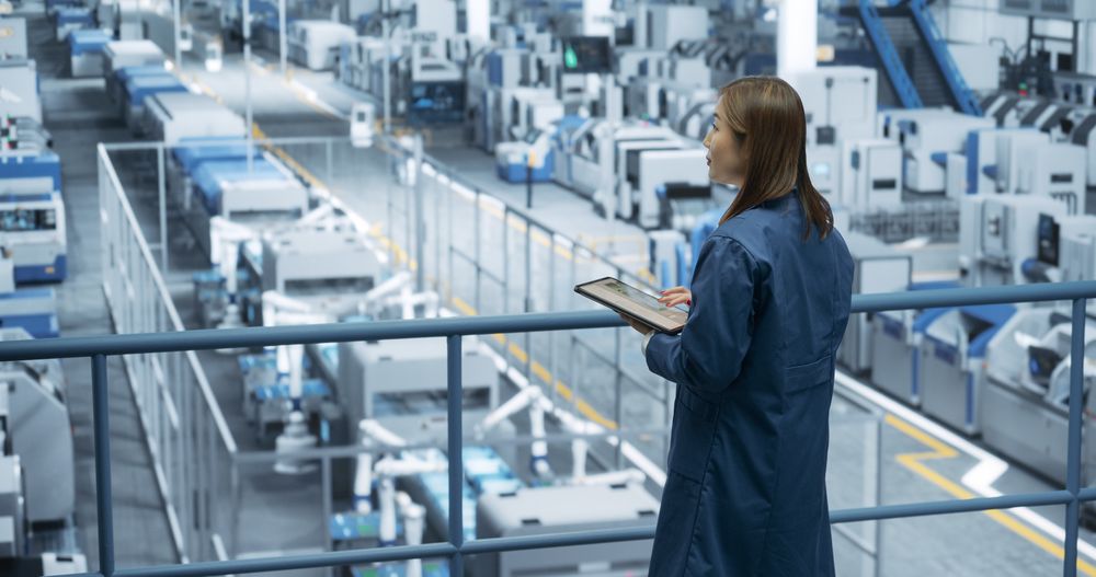 Young Asian Female Engineer Standing on a Platform, Using Tablet Computer at an Electronics Factory. Machines are Undergoing Maintenance, Specialist Monitoring the Progress Through Online Software