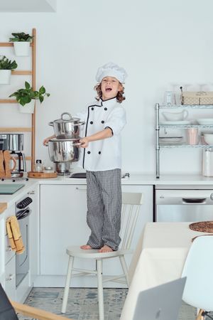 Cheerful boy in chef costume standing on chair with saucepans