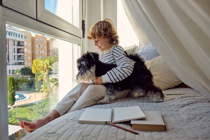 Smiling schoolboy embracing purebred dog on bed against window