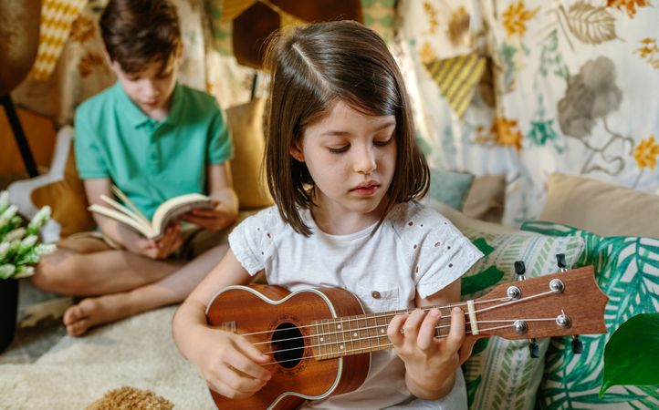 Portrait of concentrated little girl playing ukulele and boy reading book on handmade teepee at home