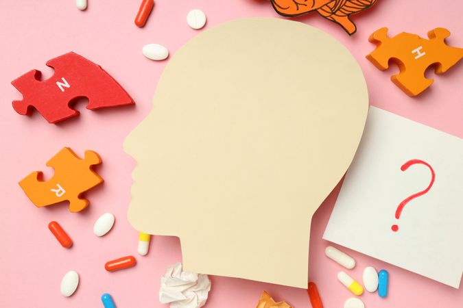 Paper cut out of side view of head with medications and puzzle pieces on pink background, close up