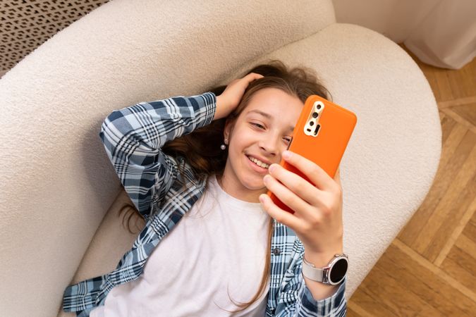 Girl smiling happily taking selfie on sofa with smartphone.