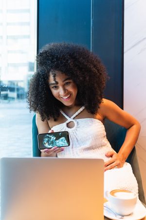 Smiling Black pregnant woman showing baby ultrasound picture to laptop