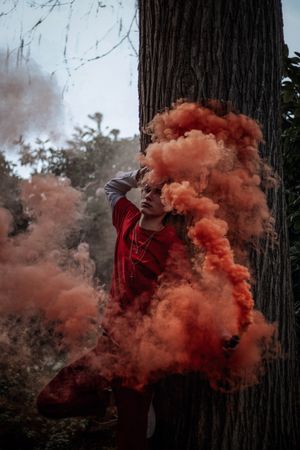 Man in red shirt and pants holding red smoke gun standing beside a tree
