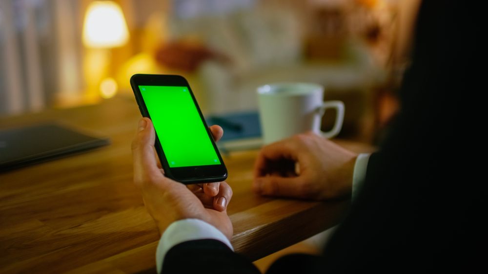 At Home Man Sits at His Desk and Uses Smartphone with Green Screen on It.