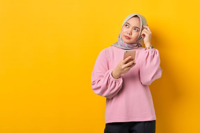 Muslim woman in headscarf contemplating something while holding smart phone and scratching head