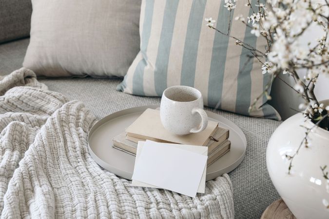 Spring breakfast scene. Blank greeting card mockup. Cup of coffee, tea on books. Round beige tray. Blossoming cherry plum tree branches in ceramic vase. Cozy linen sofa, cushions. Easter still life.