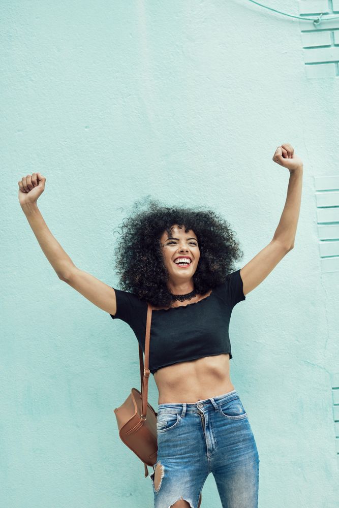 Female in casual clothes leaning on turquoise wall and smiling while raising her arms in celebration