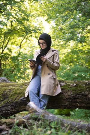 Middle Eastern woman sitting on mossy tree with book
