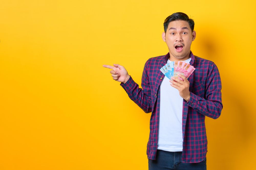 Surprised young Asian man in plaid shirt holding banknotes