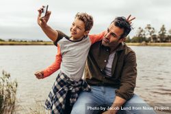 Man sitting on a tree trunk beside a lake with his son and taking a selfie 5QVeN4