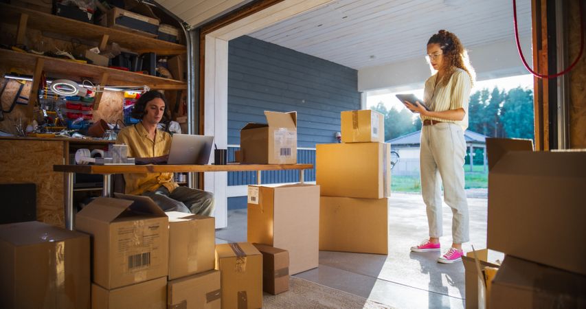Male Small Business Owner Using Laptop Computer In Home Garage To Fill Online Orders And Talking To Hispanic Female Manager.