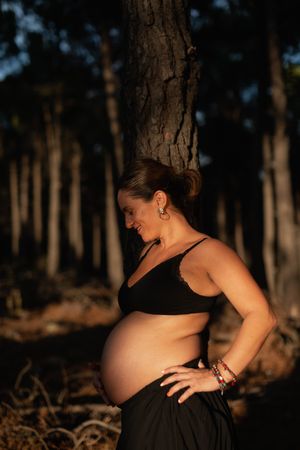 A pregnant woman holds her belly and smiles wearing underwear in the woods