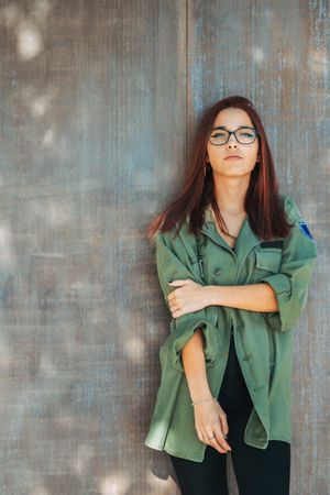Stylish confident bright woman in glasses leaning on wall and looking at camera