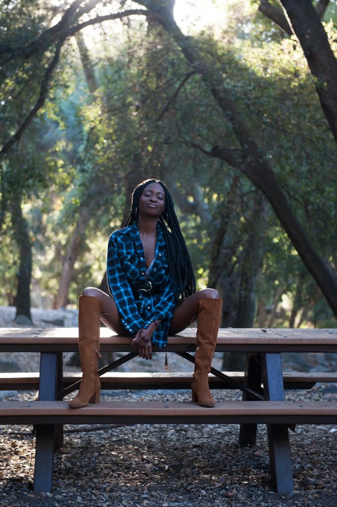Fashion photo of woman in blue flannel shirt and tall boots sitting on bench in the mountains