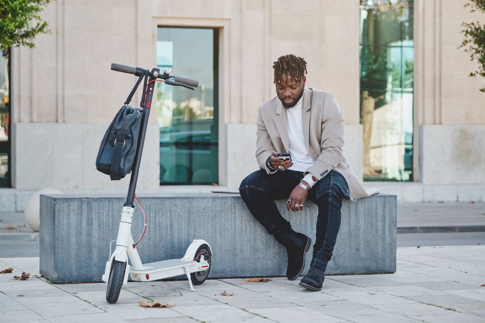 Black man sitting on bench in street with smartphone in shade o