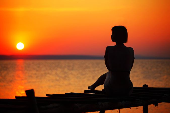 Silhouette of woman sitting on dock during sunset