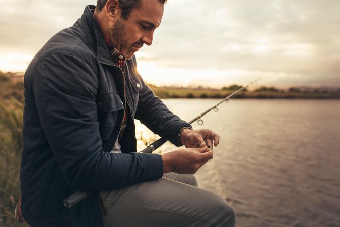 Man sitting near a lake adding bait to his fishing rod at sunrise