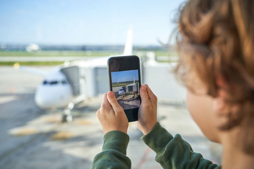 Crop kid photographing airplane on smartphone through window in