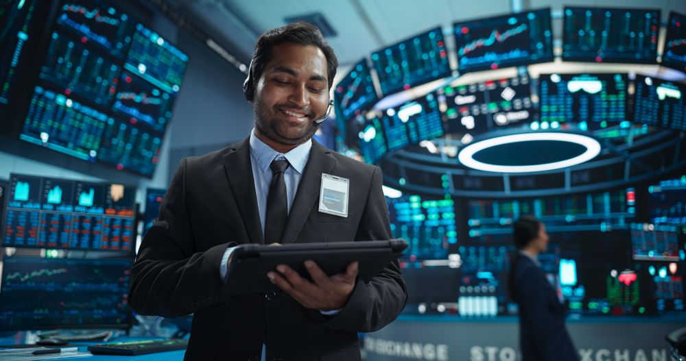 Portrait of a Young Indian Man Working in an International Stock Exchange Company: Happy Trader Using a Tablet Computer, Monitoring, Talking to a Corporate Business Partner in Headphones