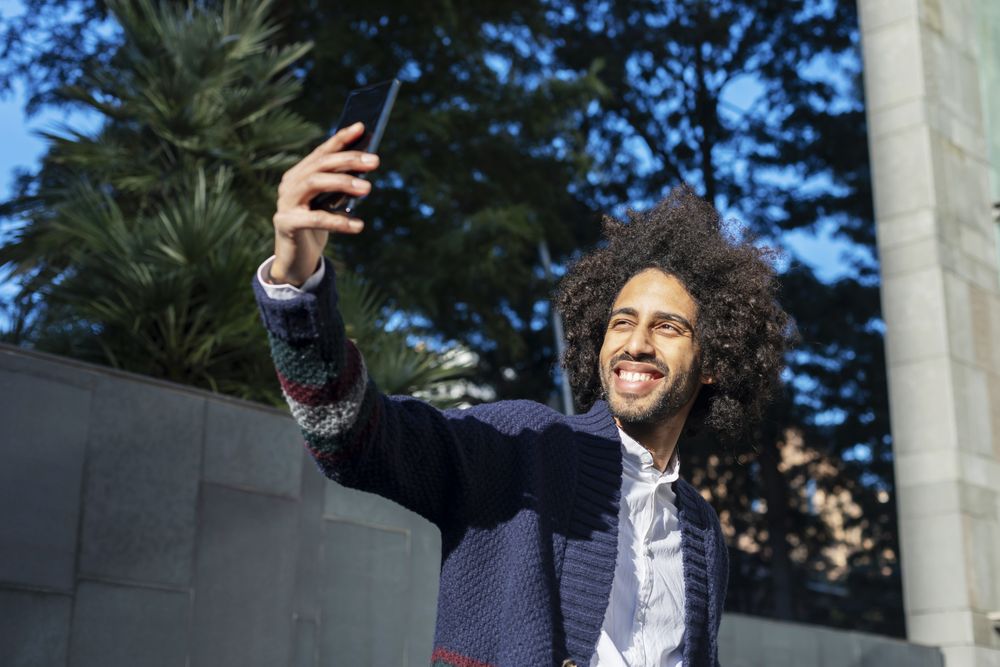 Smiling Black man taking selfie with smartphone while outside in the sun