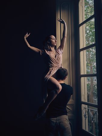 Ballerina on shoulder of faceless dancer near window