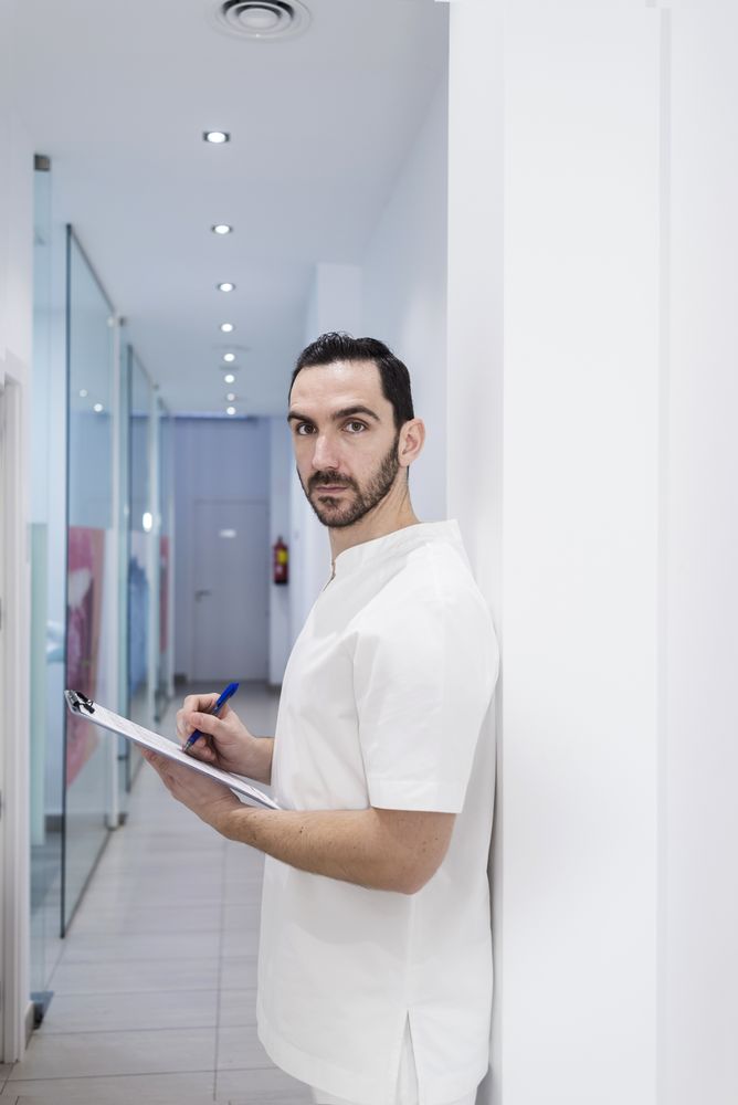 Portrait of a bearded doctor holding a clipboard while leaning on wall in clinic corridor